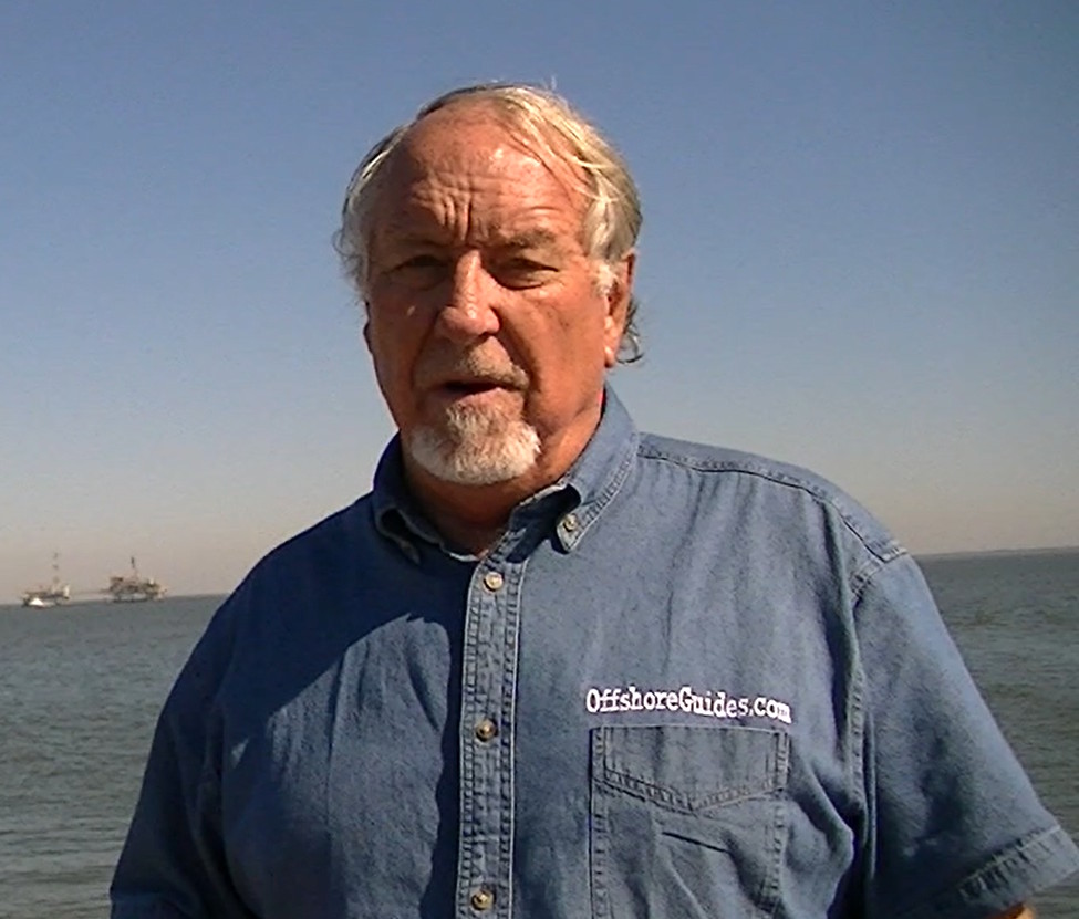 Man wearing an OffshoreGuides.com shirt standing on a boat at sea with offshore platforms in the background